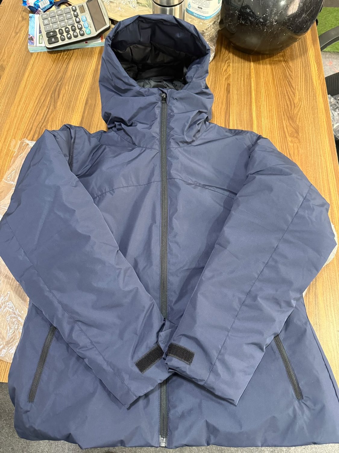 A blue grey hooded jacket laid on a wooden table beside a calculator and a flask