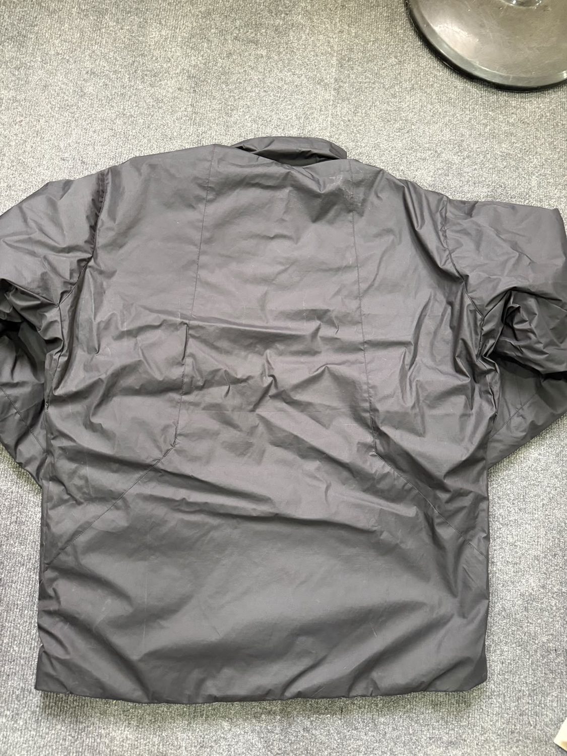 The plain back panel of a grey shell jacket laid flat on a grey carpet floor